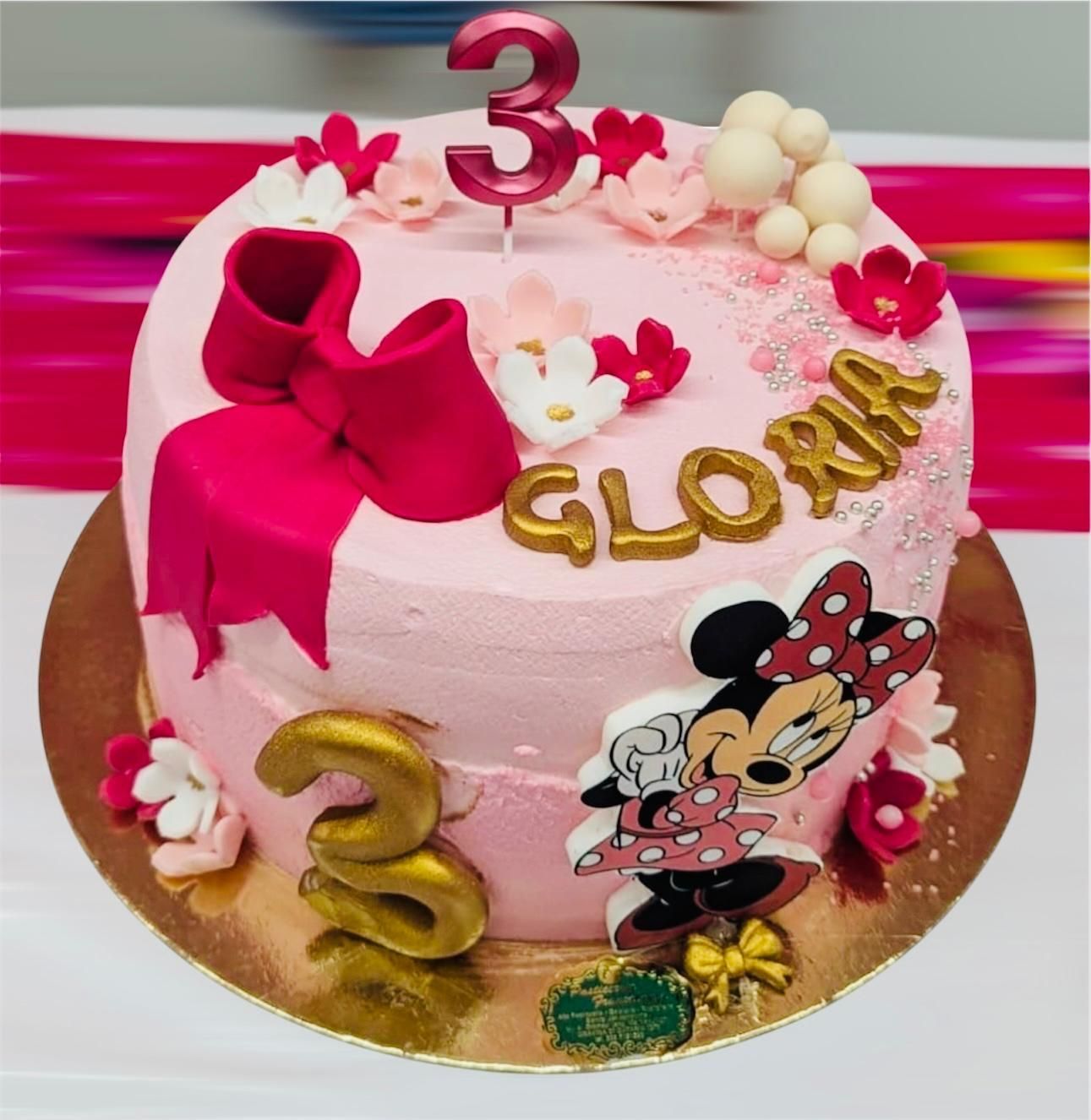 Torta di compleanno rosa a tema Minnie per una bambina di tre anni di nome Gloria, decorata con un fiocco, fiori e numeri dorati.