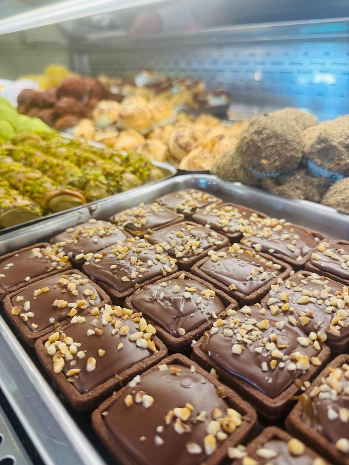 Vassoio di crostatine al cioccolato guarnite con noci tritate, esposte in una vetrina da pasticceria insieme ad altri dolci.