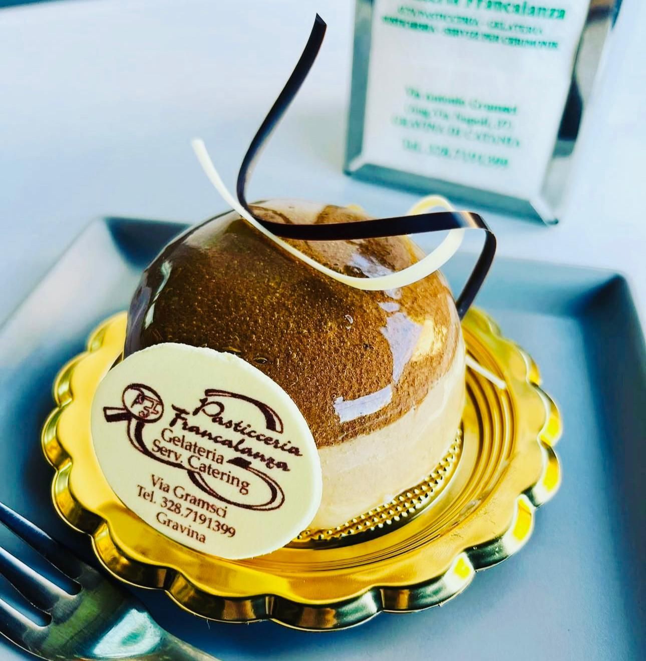 Dessert su un piatto d'oro: torta a forma di cupola con glassa, spirali di cioccolato, logo e una forchetta.