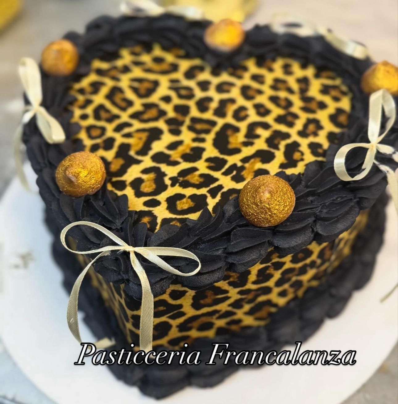 Torta a forma di cuore con motivo leopardato, bordo glassato nero, dettagli dorati e fiocchi di nastro.