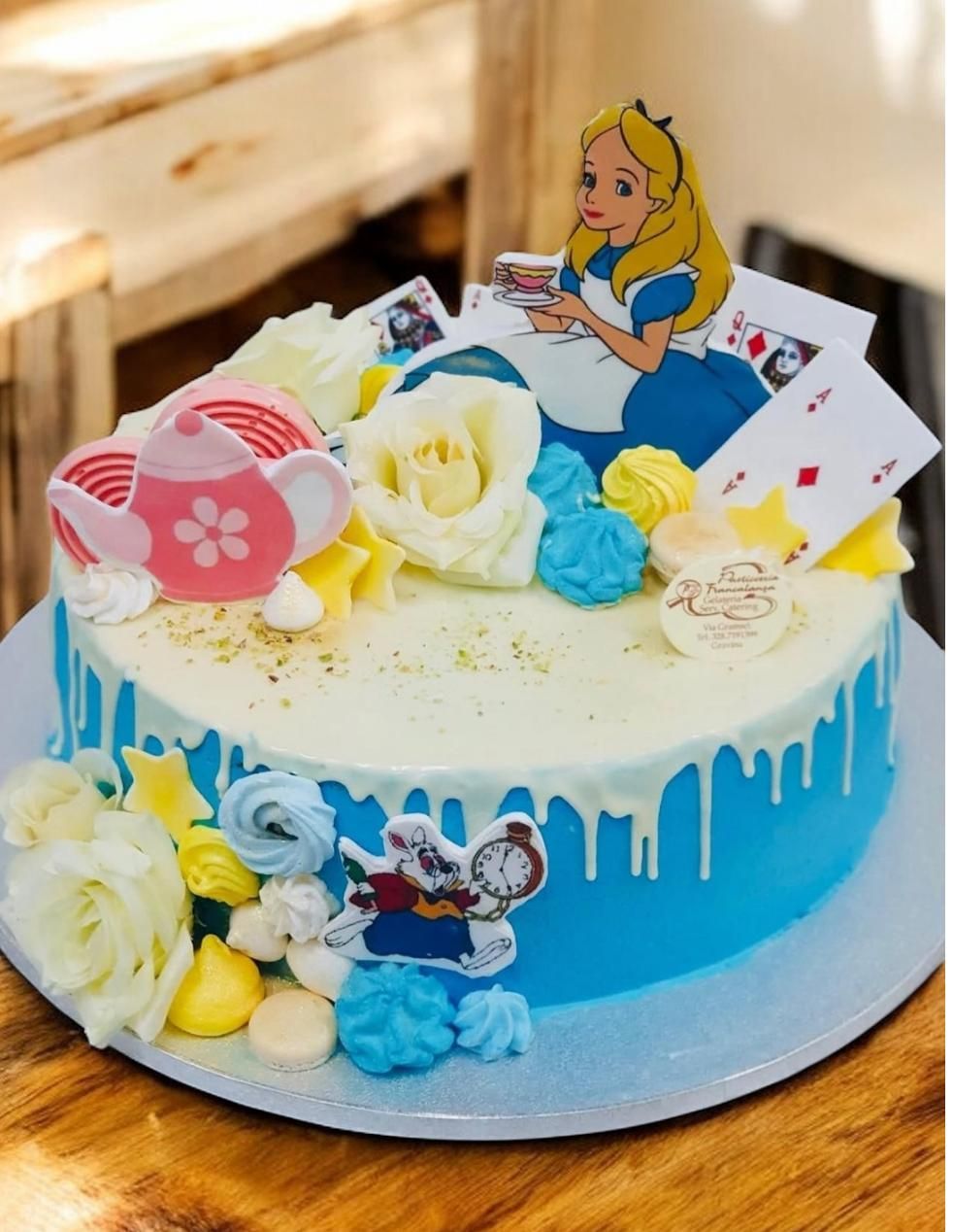 Torta a tema Alice nel Paese delle Meraviglie con glassa blu, gocce bianche e decorazioni raffiguranti personaggi come Alice e le carte da gioco.
