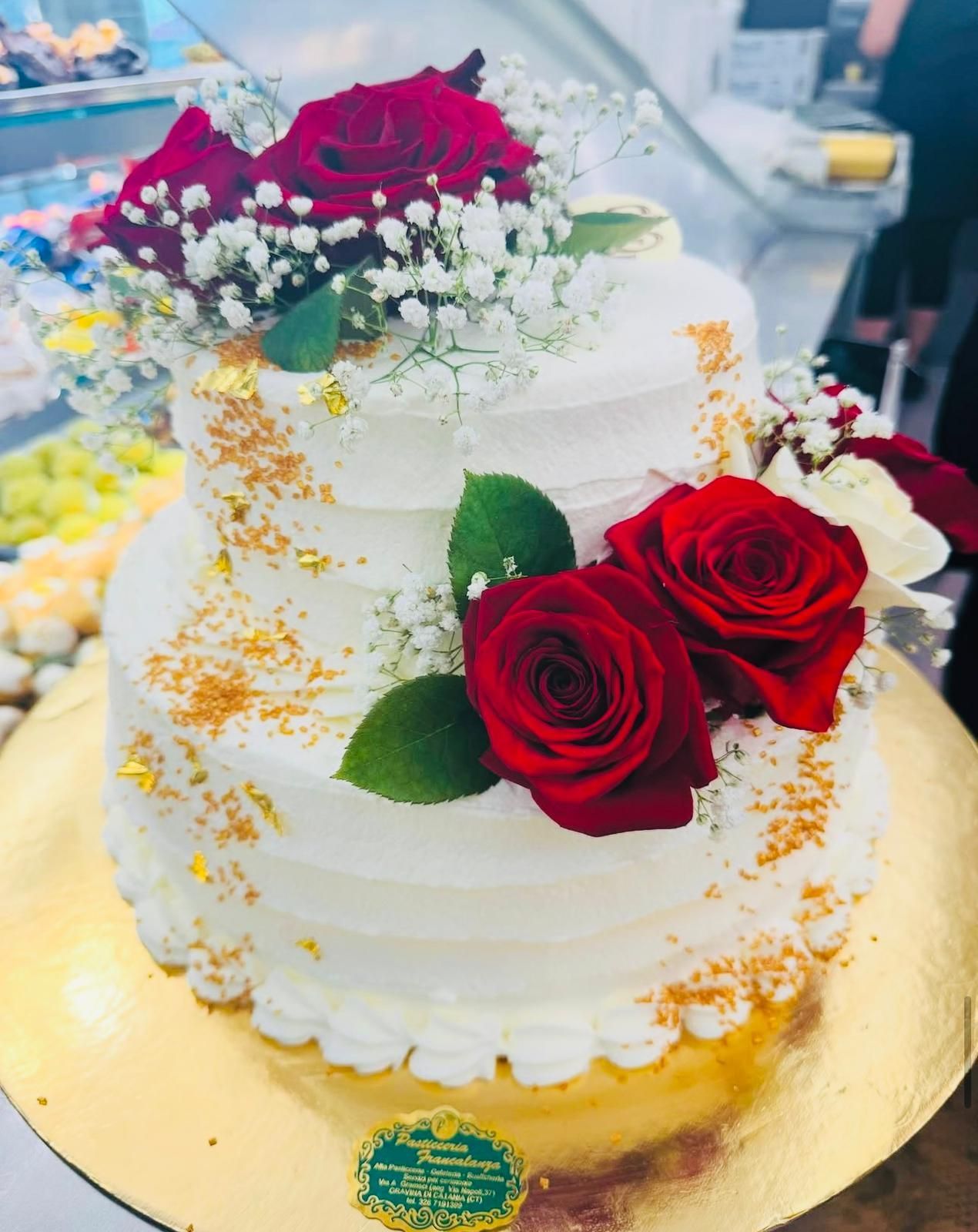 Torta bianca a due piani, decorata con rose rosse, gypsophila e pagliuzze dorate, su una base dorata.