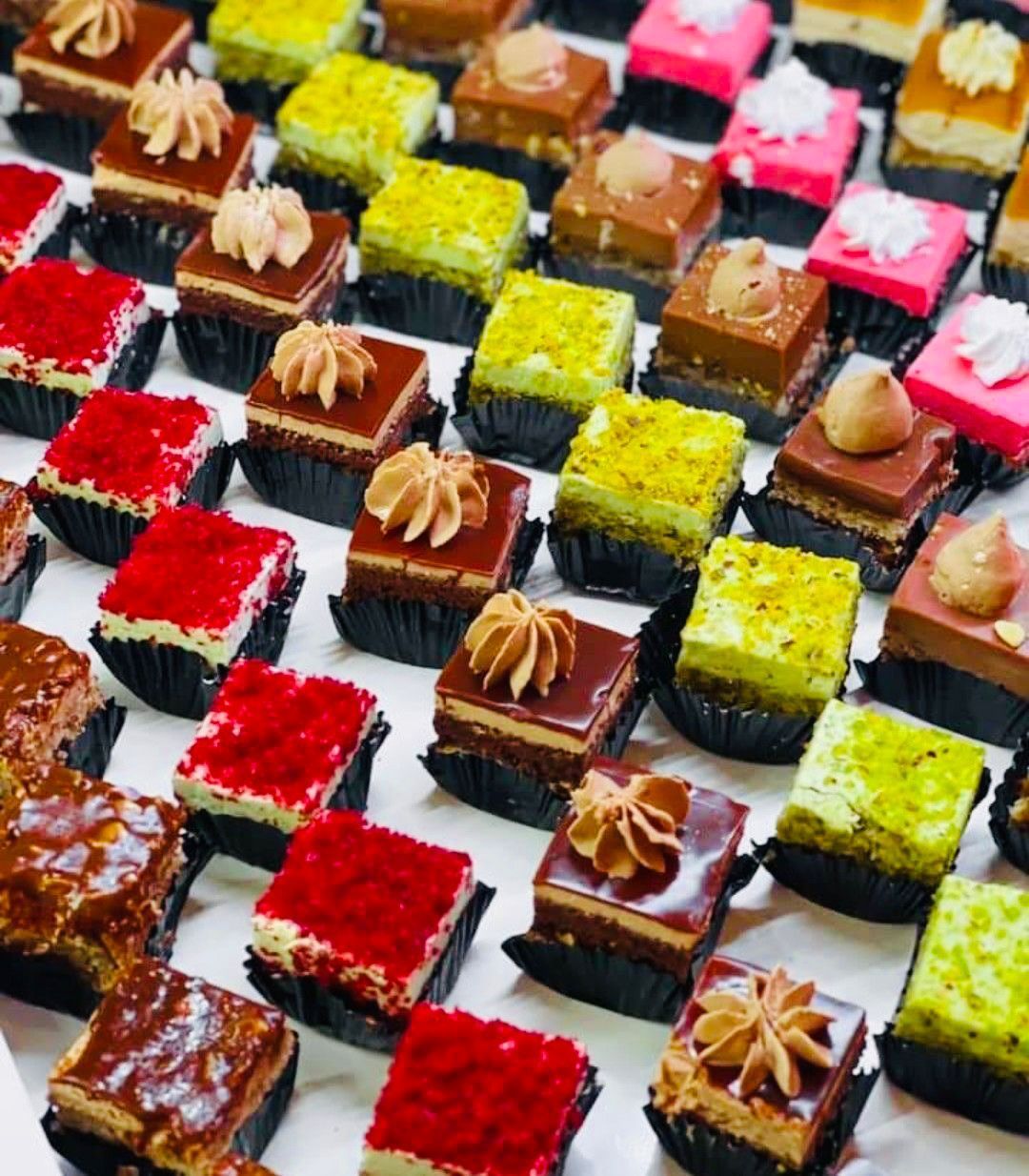 File di mini-torte colorate e quadrate in pirottini di carta neri. I gusti includono cioccolato, rosso, rosa e pistacchio, decorati con glassa.