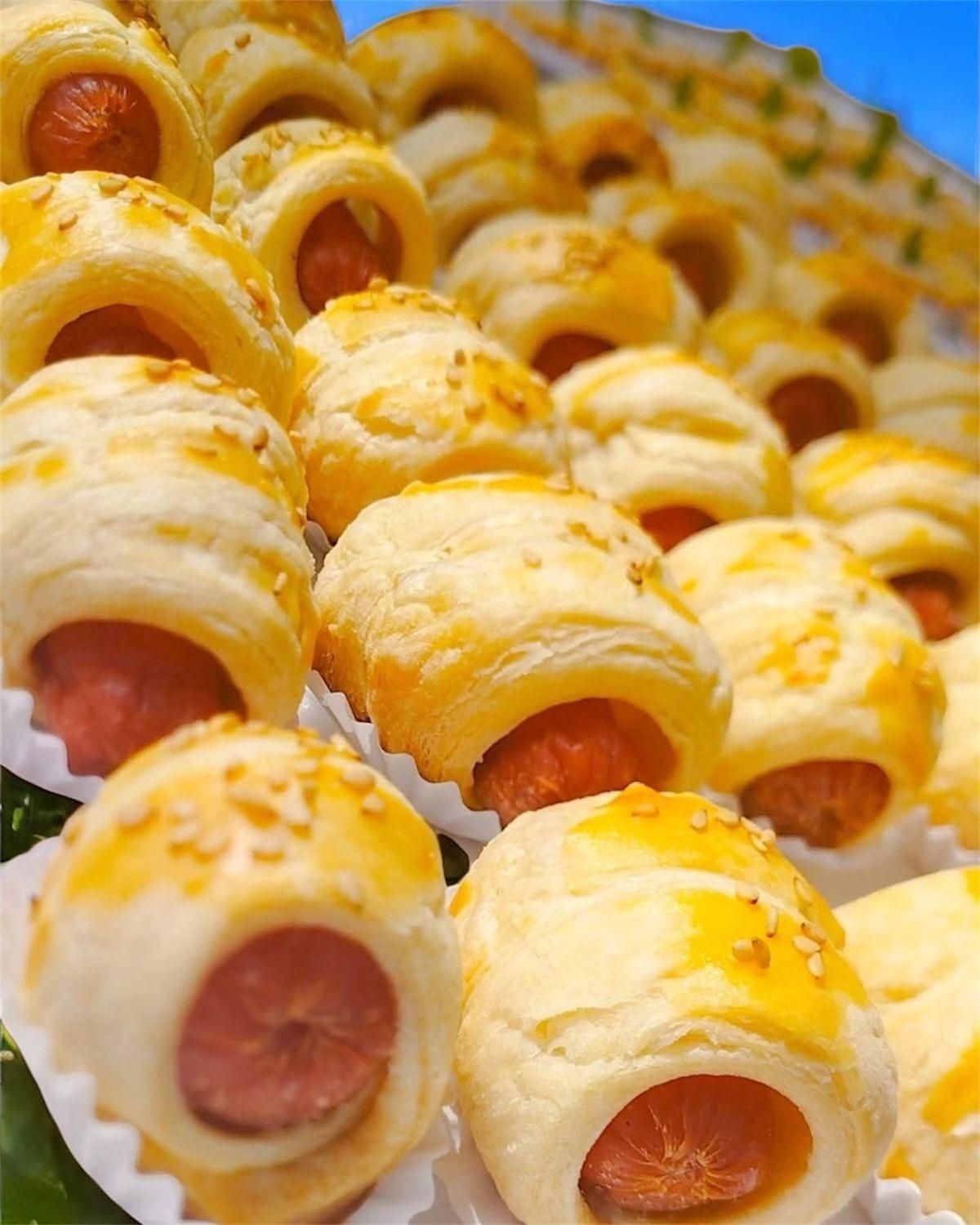 Primo piano di involtini di salsiccia; pasta sfoglia dorata avvolta attorno a hot dog, alcuni con semi di sesamo.