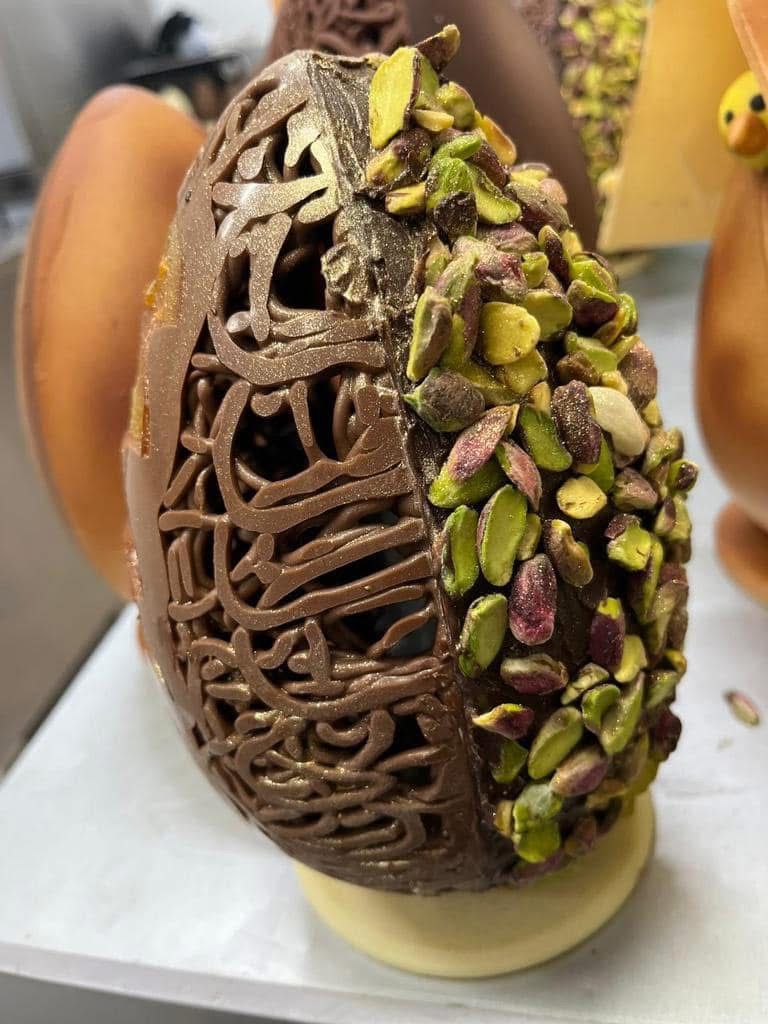 Uovo di Pasqua di cioccolato, metà intagliato e metà ricoperto di pistacchi, su base bianca.