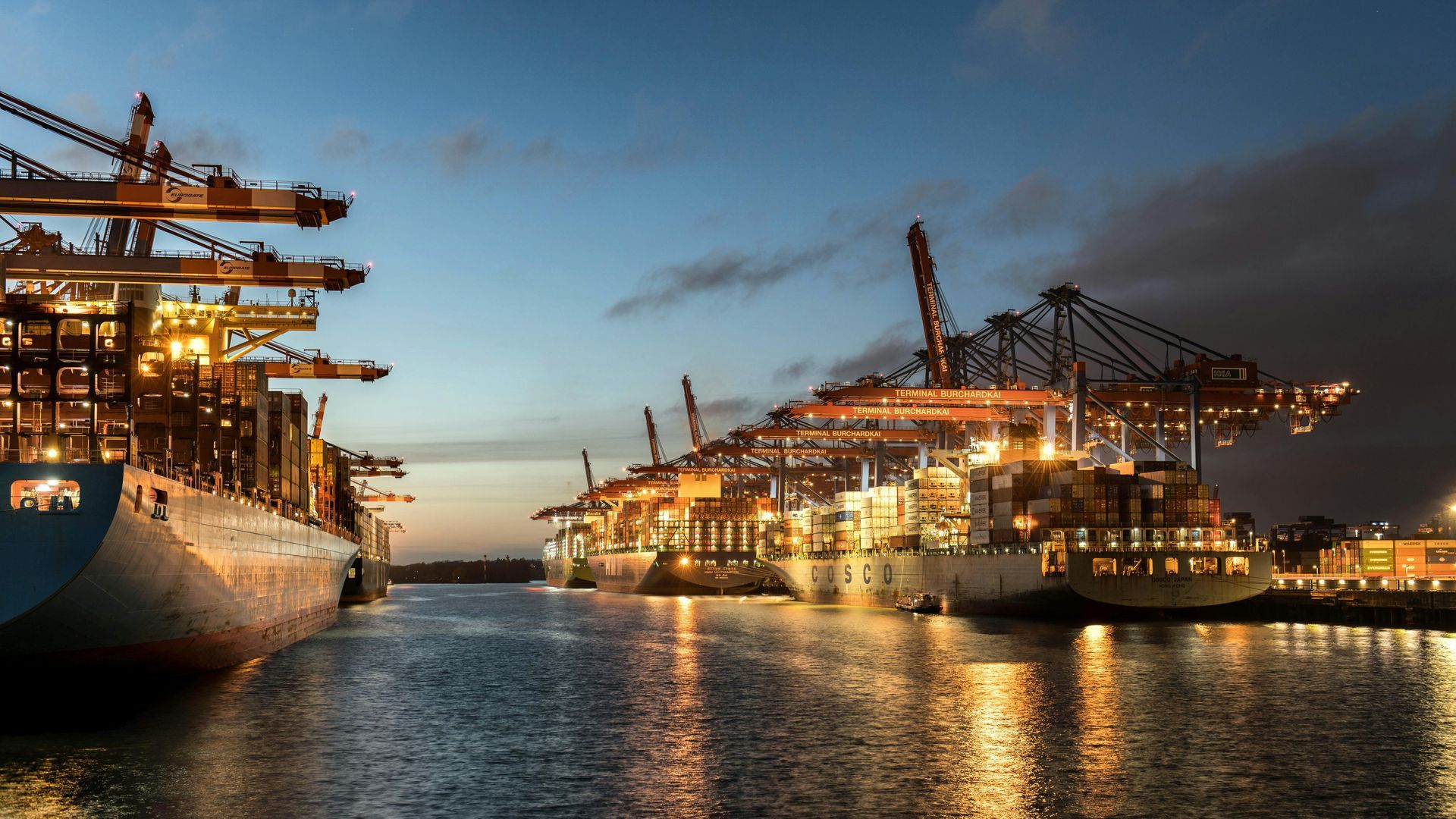 Containerschepen in een helder verlichte haven bij schemering, met kranen die lading laden.