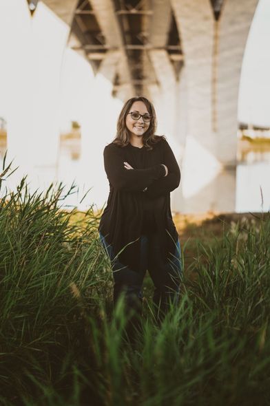 A woman is standing in the grass under a bridge with her arms crossed.