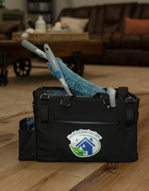 Black cleaning caddy with supplies on wood floor, logo on front, duster and other tools visible, living room background.