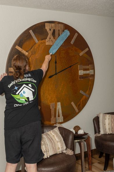 Person dusting a large wall clock with a blue duster, in a living room.