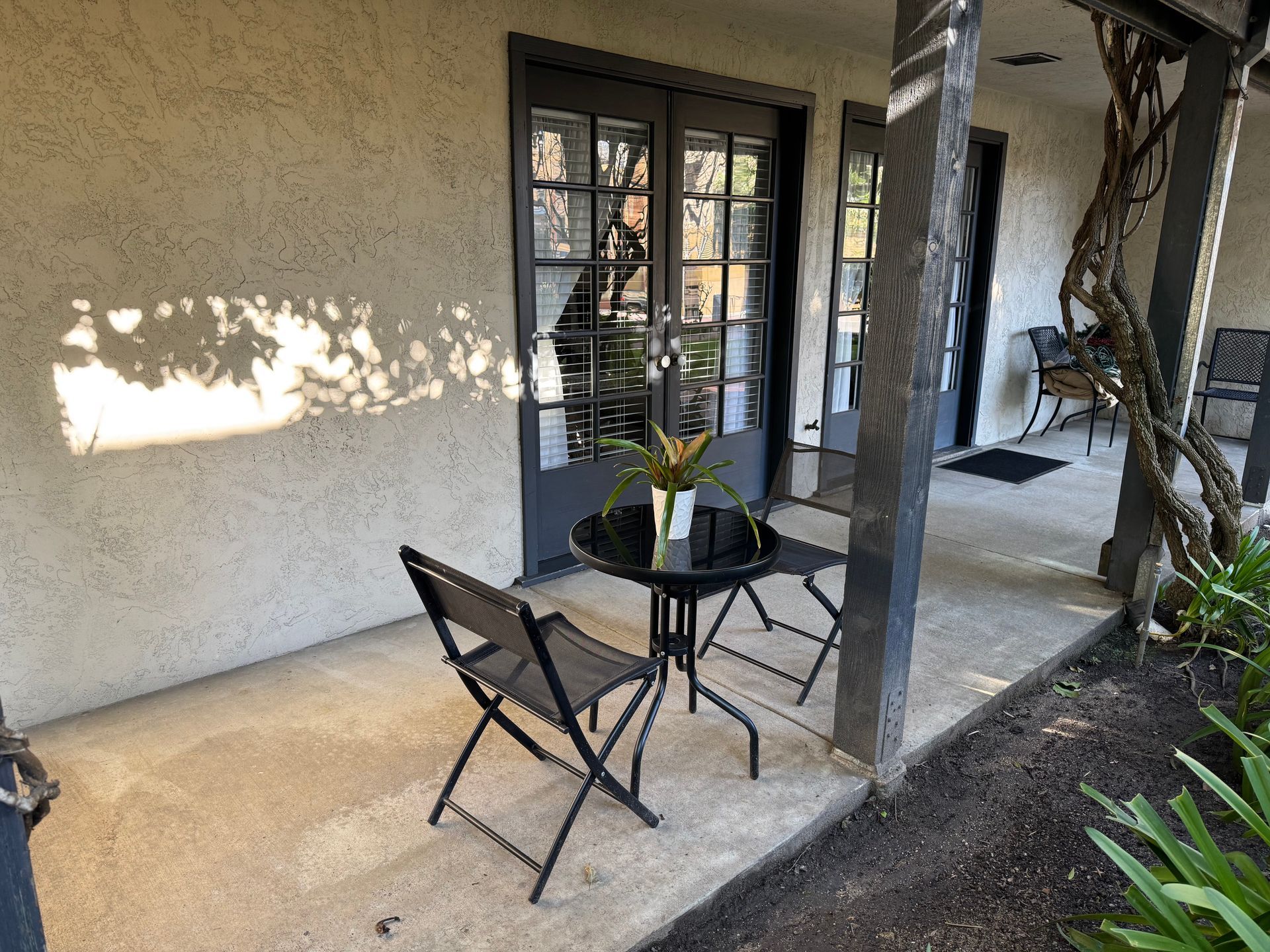 Outdoor seating area with table, chairs, and French doors. Beige stucco wall, dark gray trim.