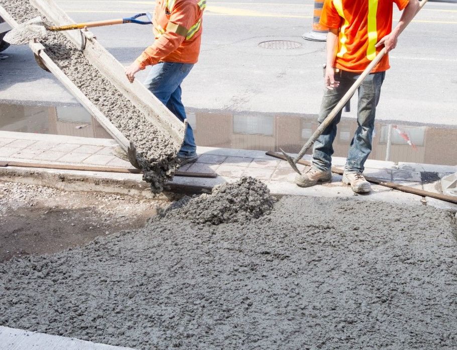 Construction workers pouring wet concrete onto a roadway from a chute, wearing orange safety vests.