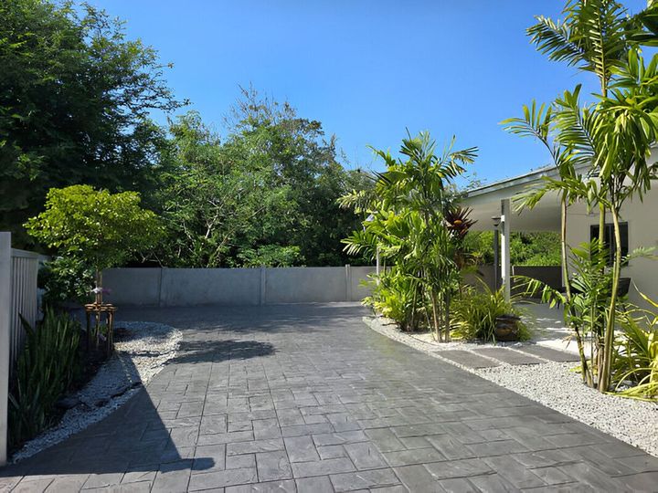 Sunlit paved driveway beside a white house, lined with tropical plants and a gray wall