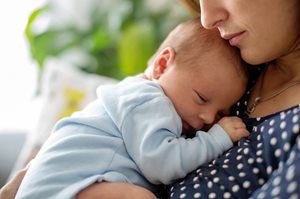 Woman holding sleeping baby in blue outfit, with a soft green background