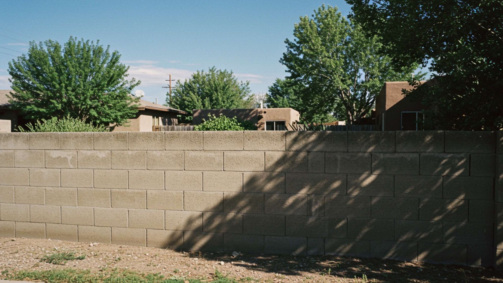 Concrete block wall with trees and houses in the background under a blue sky.