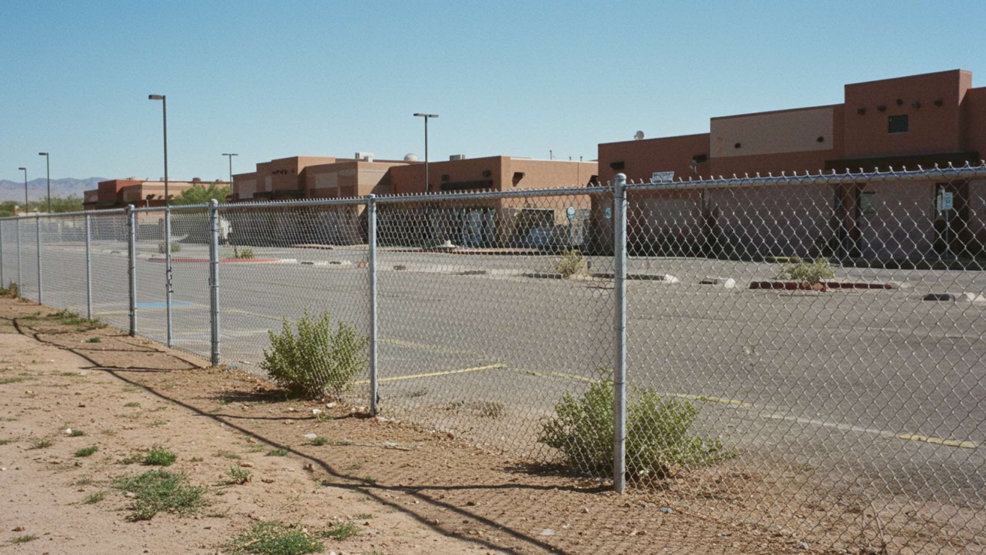 Chain-link fence with buildings in the background and sparse vegetation in a desert setting.