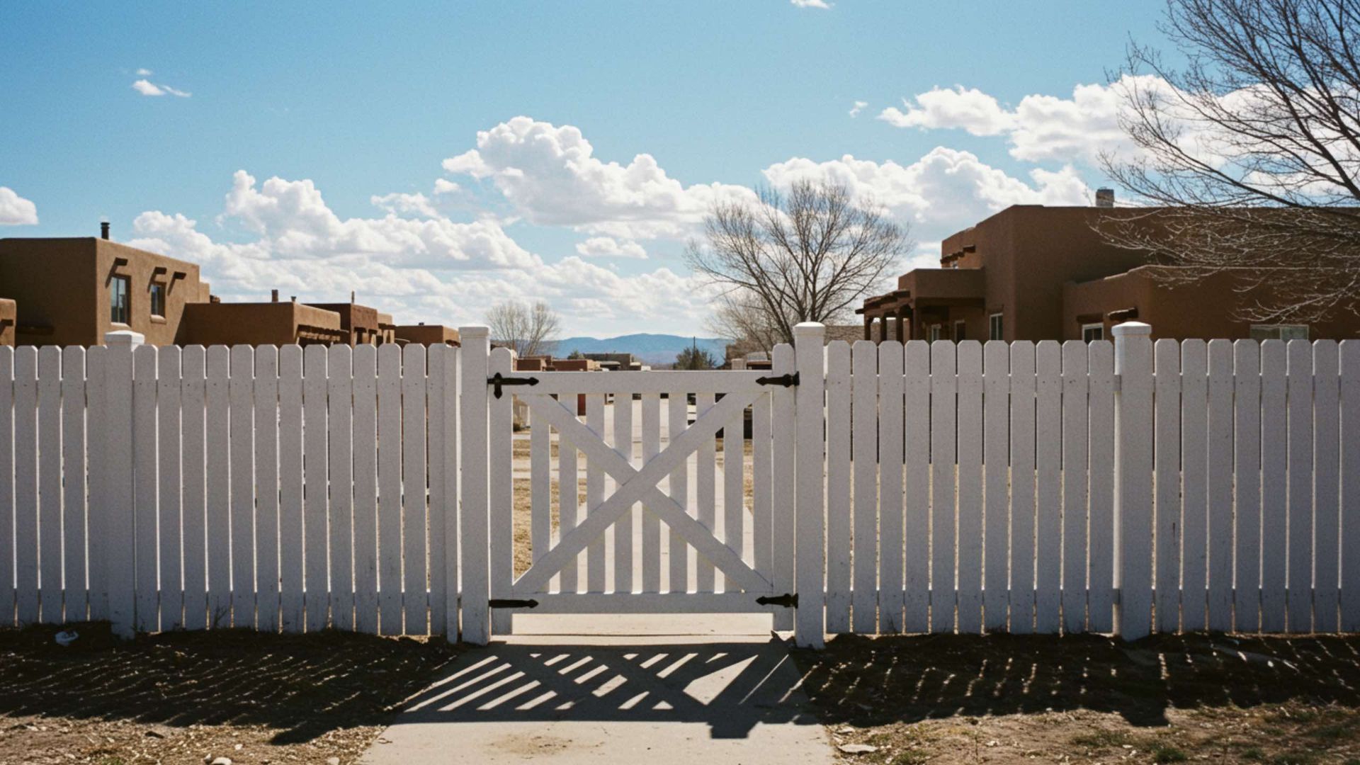 A white picket fence with a centered gate stands before tan stucco houses under a blue sky with white clouds.