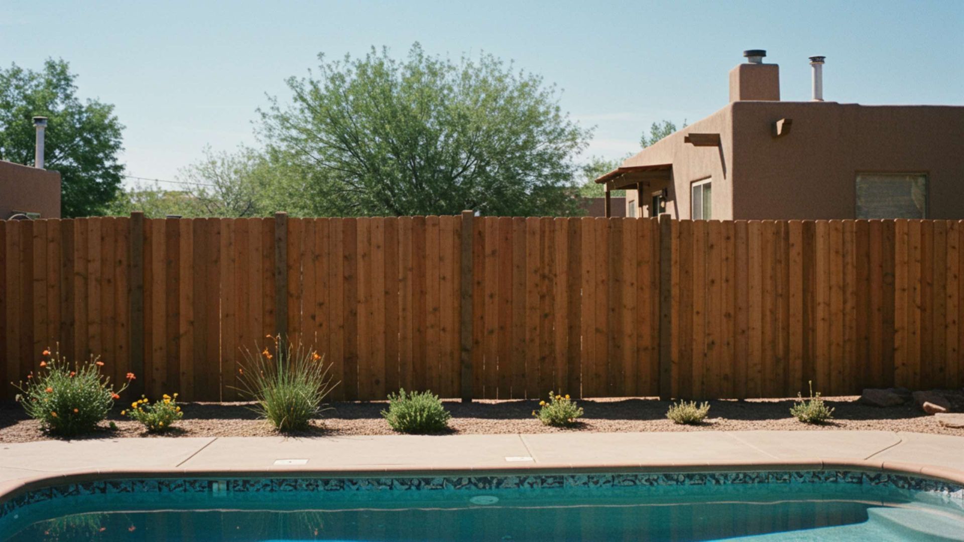 A swimming pool in a backyard with a wooden privacy fence, sparse desert landscaping, and a tan house in the background.