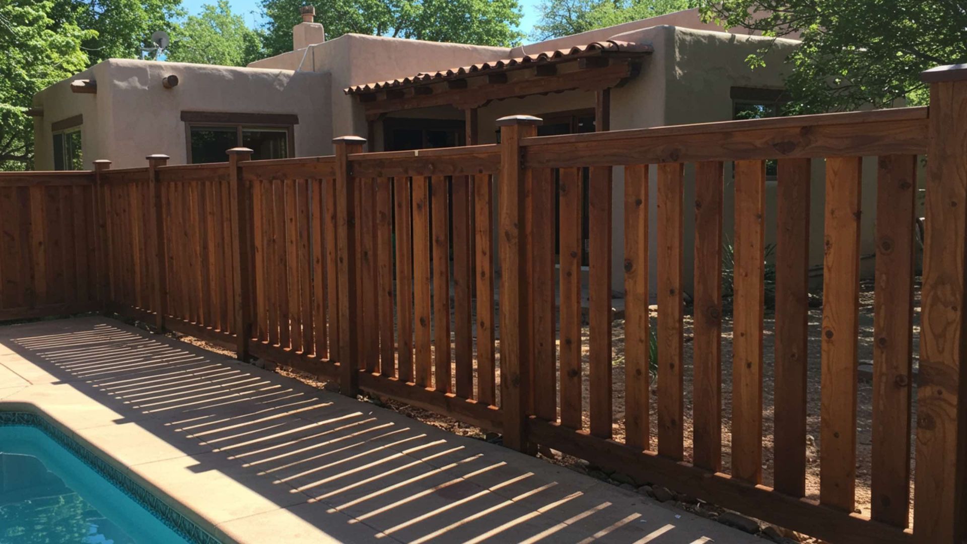 A tall, stained-wood privacy fence borders a swimming pool and patio in front of a tan stucco house.