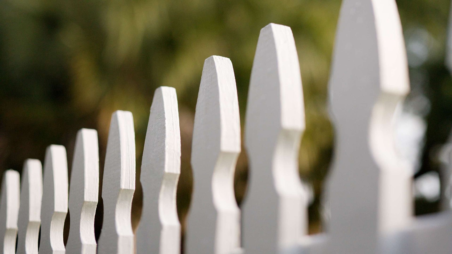 A close up of a white picket fence with trees in the background.