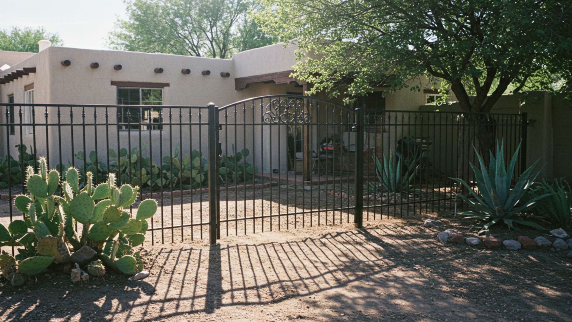 A black metal gate in front of a tan adobe-style house with cacti and a tree.