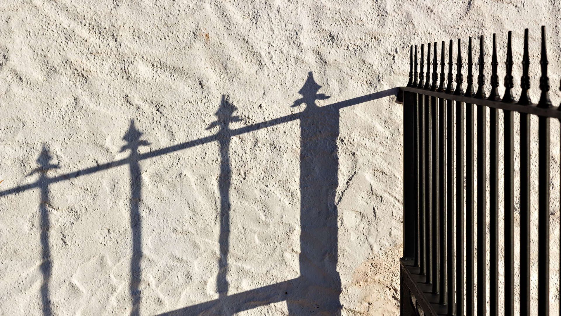 A shadow of a fence is cast on a white wall.