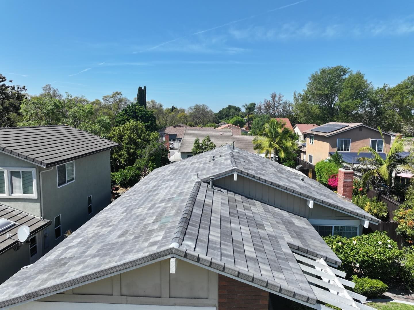 An aerial view of a dark metal-roofed house surrounded by trees, with a concrete driveway leading to the entrance.