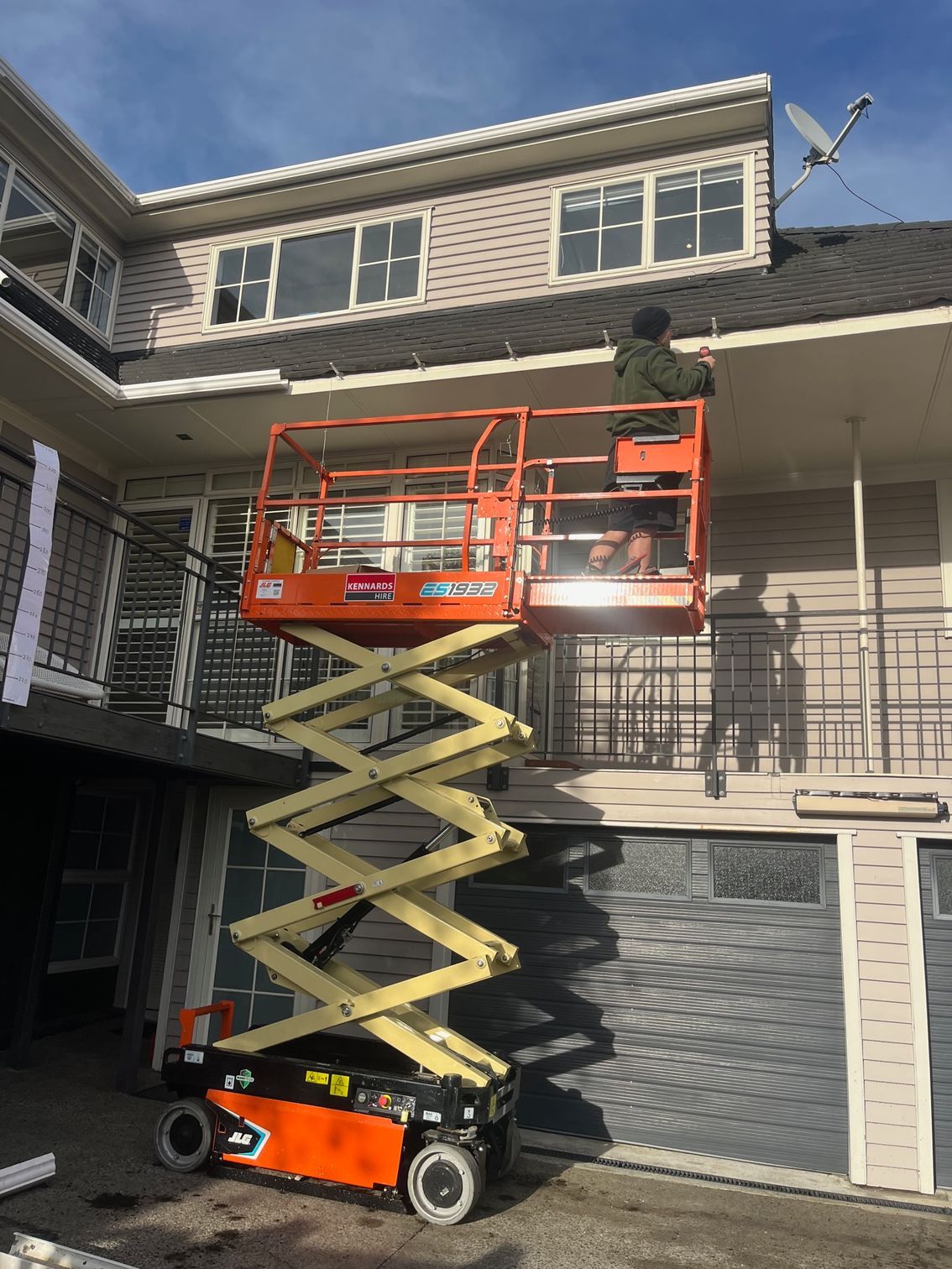 Man on an orange scissor lift works on a house's trim. The house is two-story with a gray exterior and garage.