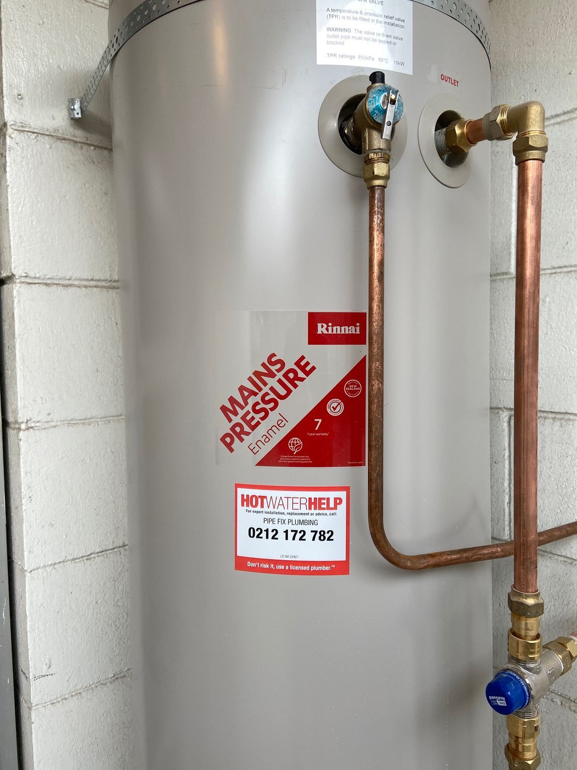 A gray mains pressure hot water heater, with copper pipes, mounted against a brick wall.