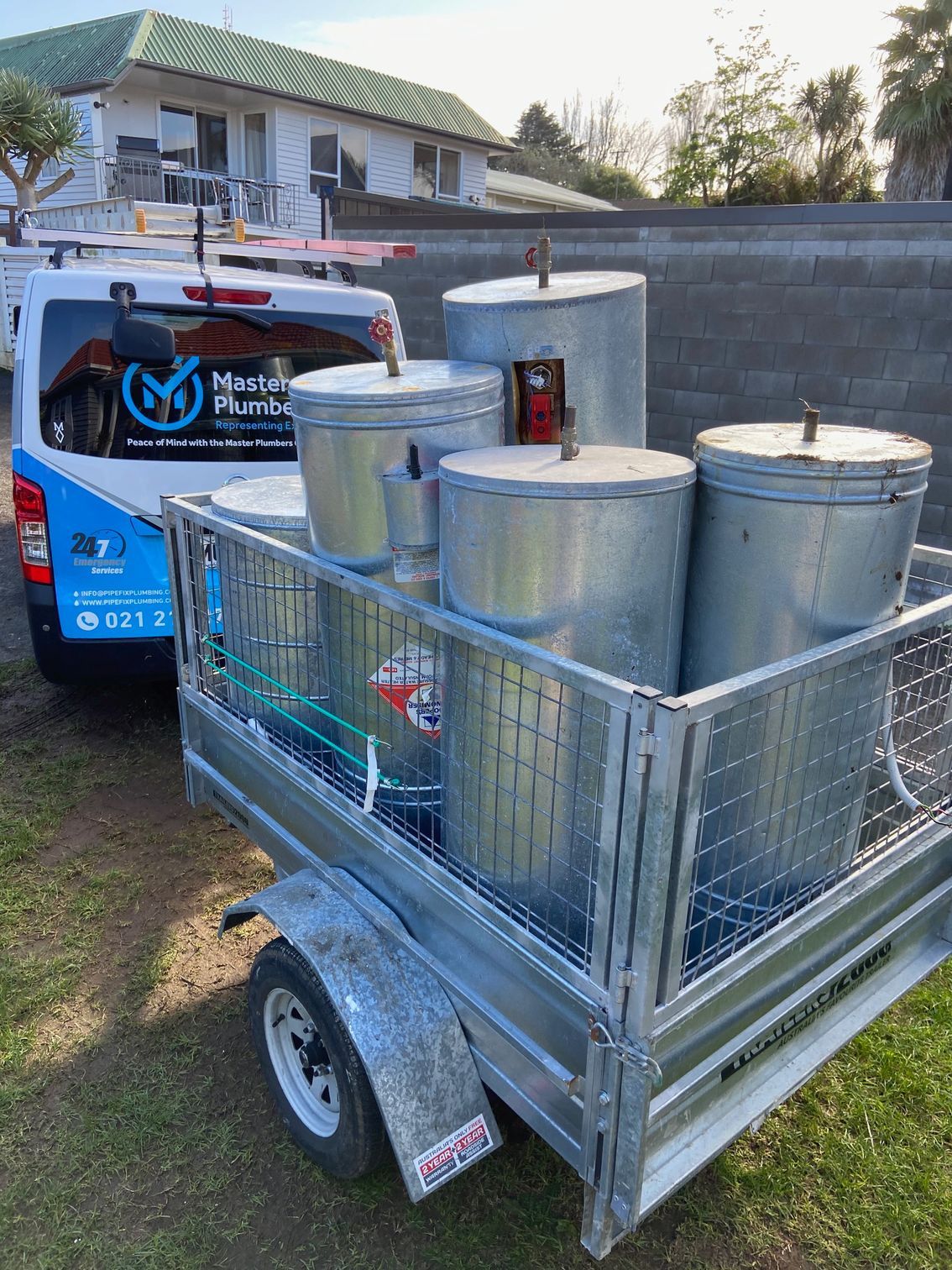 A trailer loaded with galvanized water heaters behind a plumbing service van on grass.