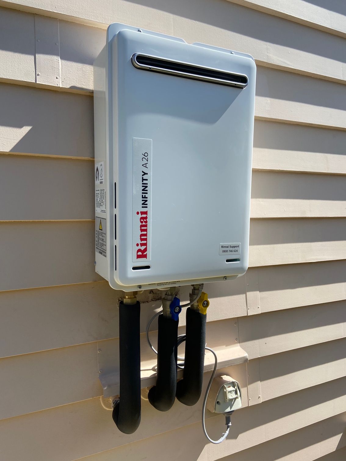 White tankless water heater on a beige siding wall, with black pipes and gas lines.