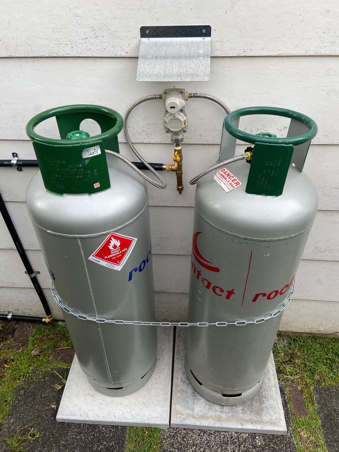 Two silver propane tanks connected to a regulator on a white wall.