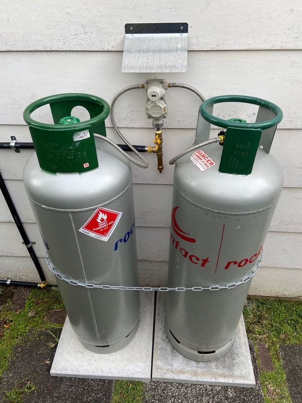 Two silver propane tanks connected to a regulator on a white wall.