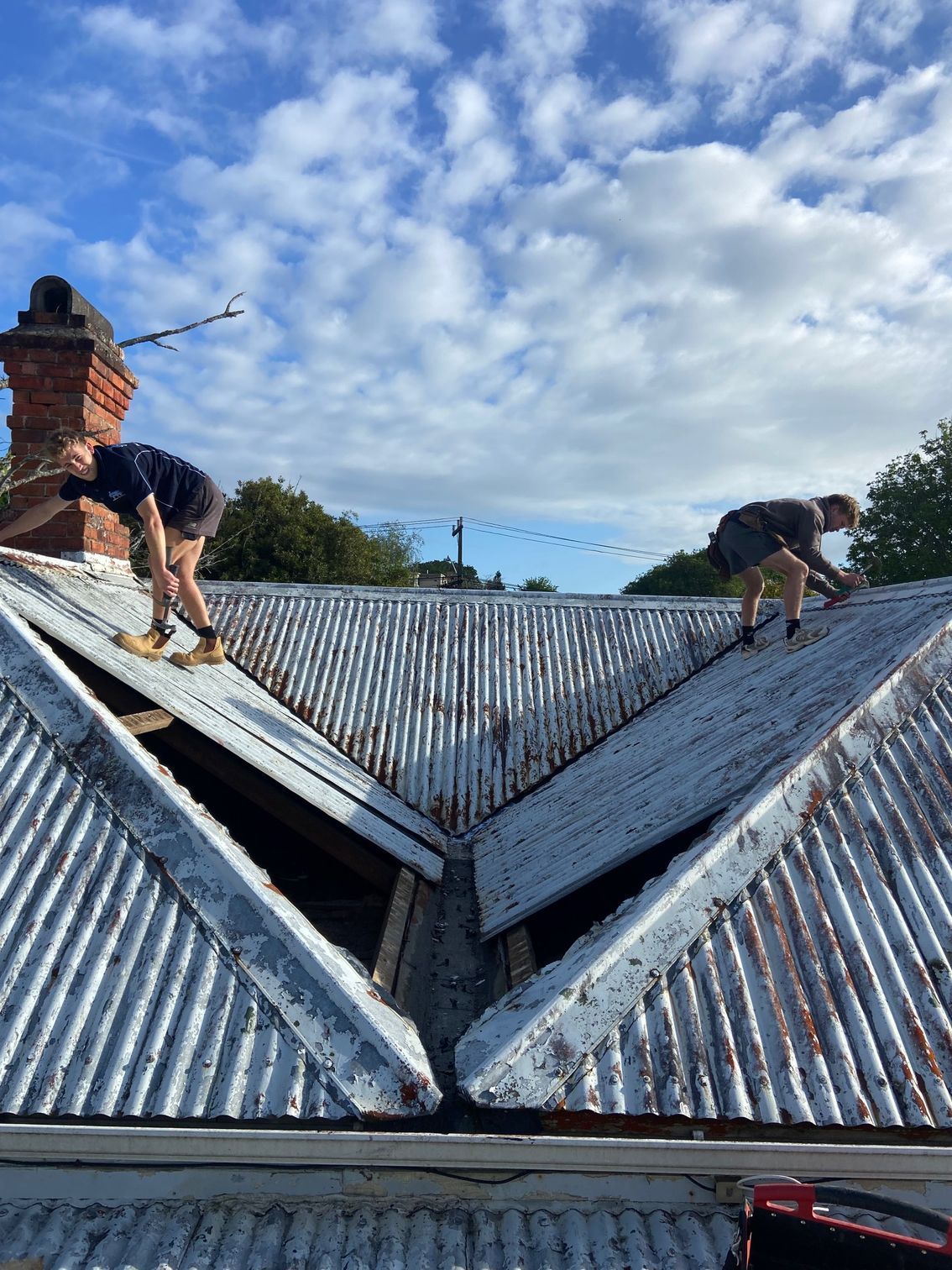 Two people working on a corrugated metal roof on a sunny day, with blue sky and chimney visible.