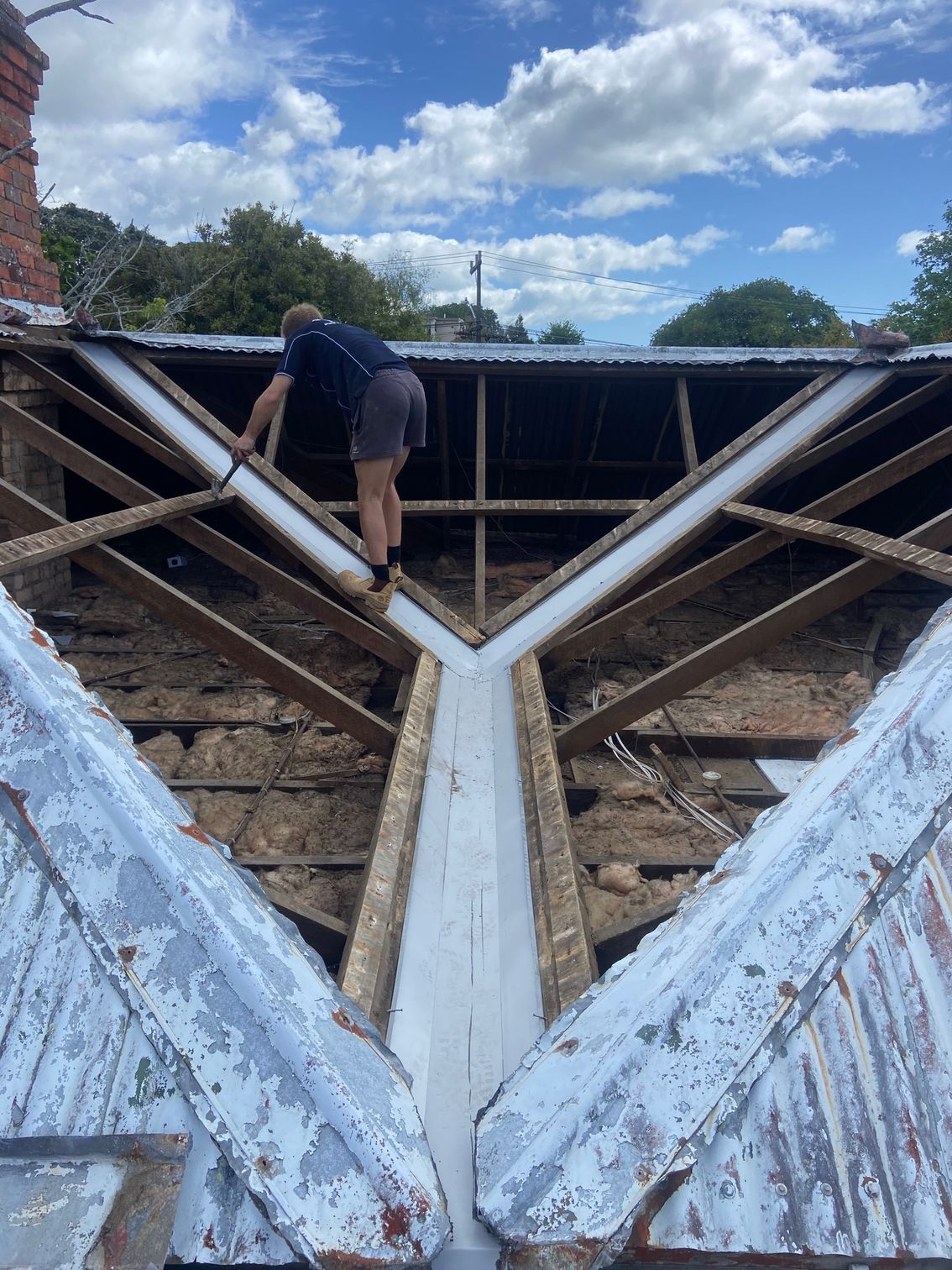 Person on roof removing shingles, blue sky. Old beams and metal trough in between roof sections.