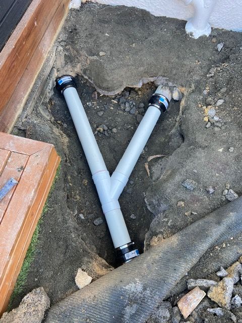 Gray plumbing pipes in a Y-shape, set in a dirt hole next to a wooden structure.