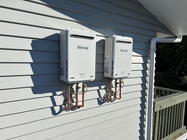 Two white Rinnai water heaters mounted on a gray house exterior wall with copper pipes and a gutter.