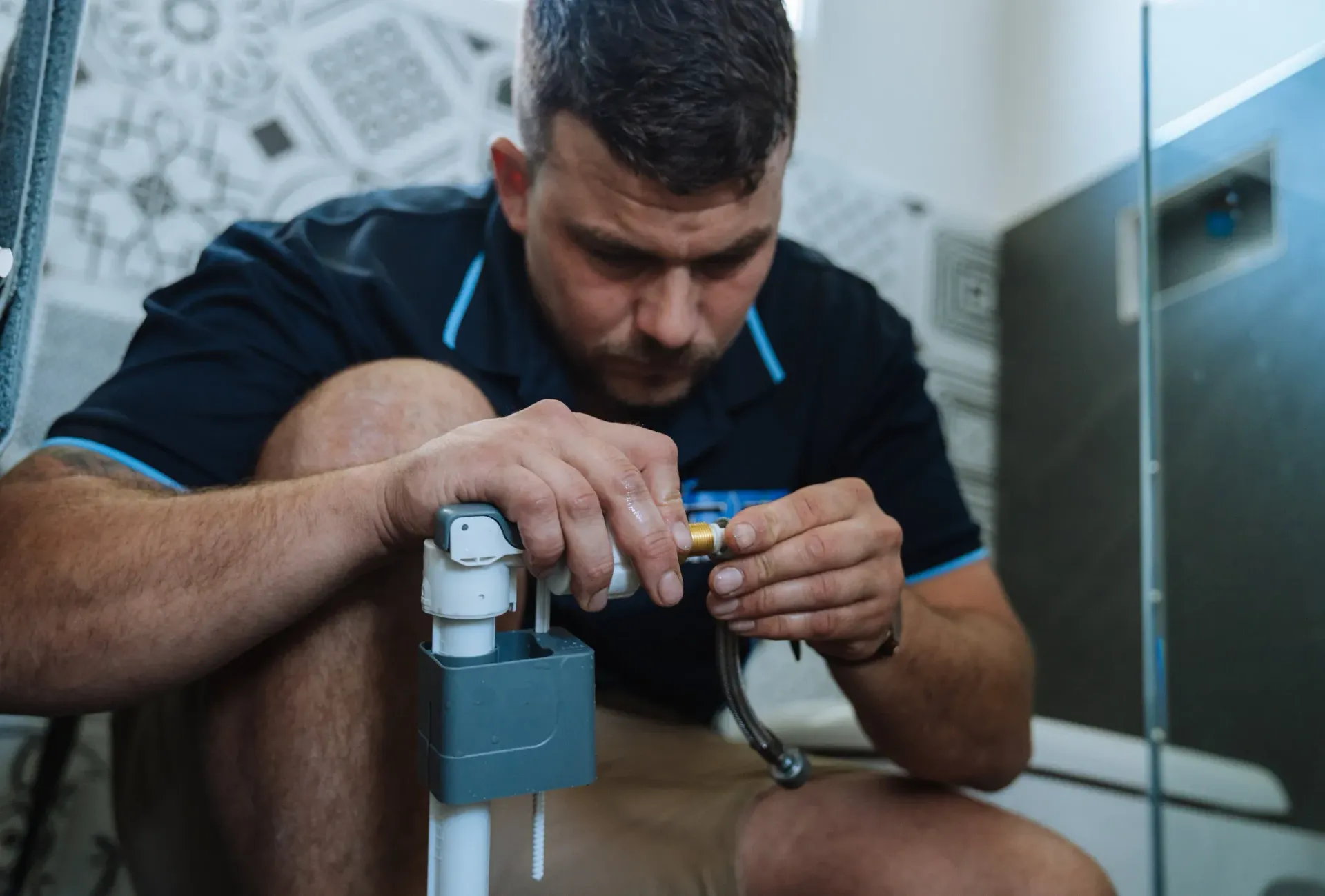 Plumber kneeling in bathroom, repairing plumbing fixture; blue and white tones.