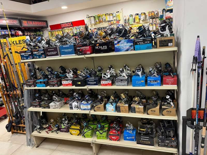 Su uno scaffale di un negozio al dettaglio sono esposti numerosi mulinelli da pesca confezionati in vari colori e modelli, con le canne da pesca in un angolo.