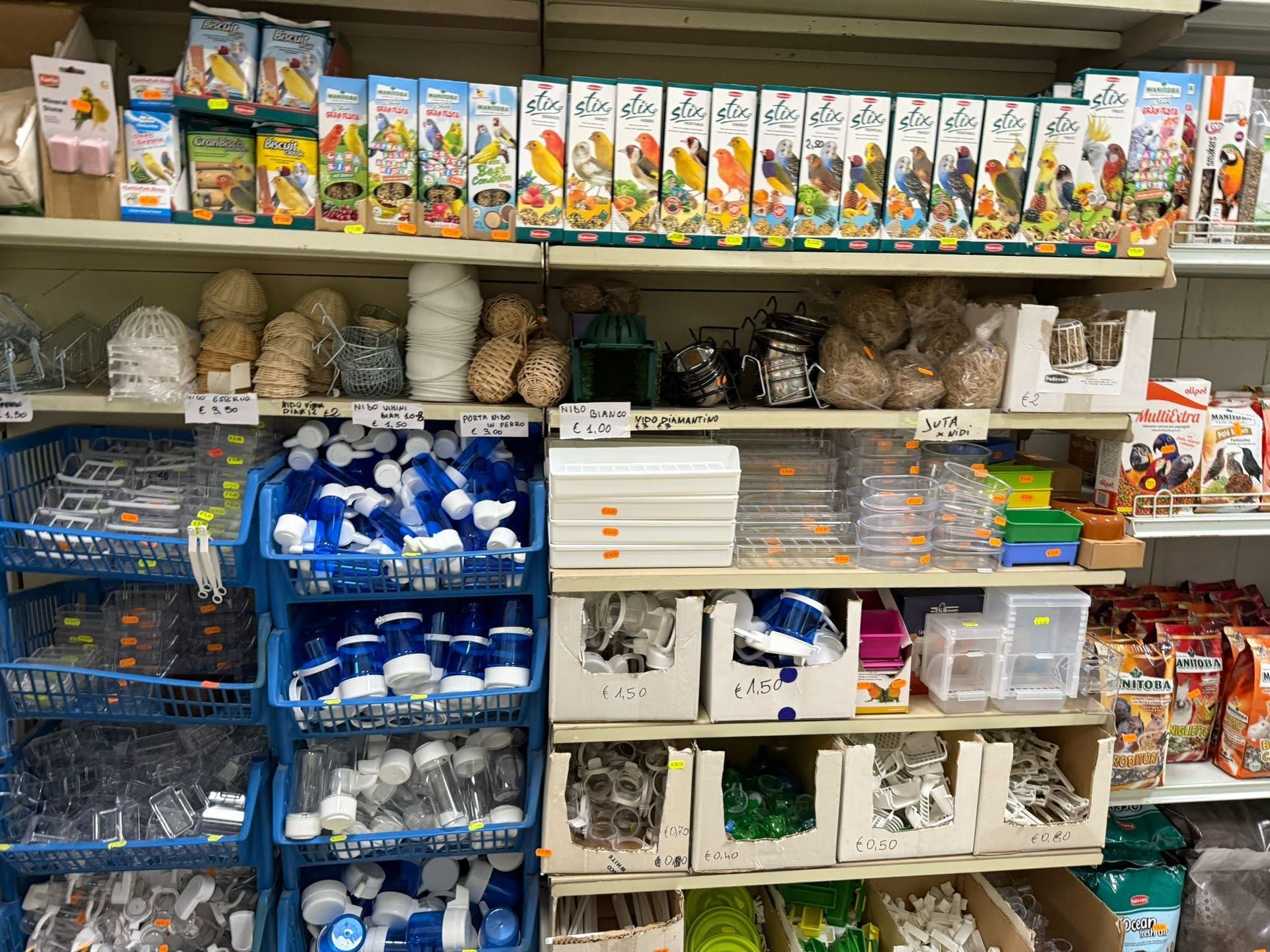 Scaffali del negozio pieni di scatole di cibo per uccelli domestici, giocattoli intrecciati e vari contenitori e mangiatoie in plastica.