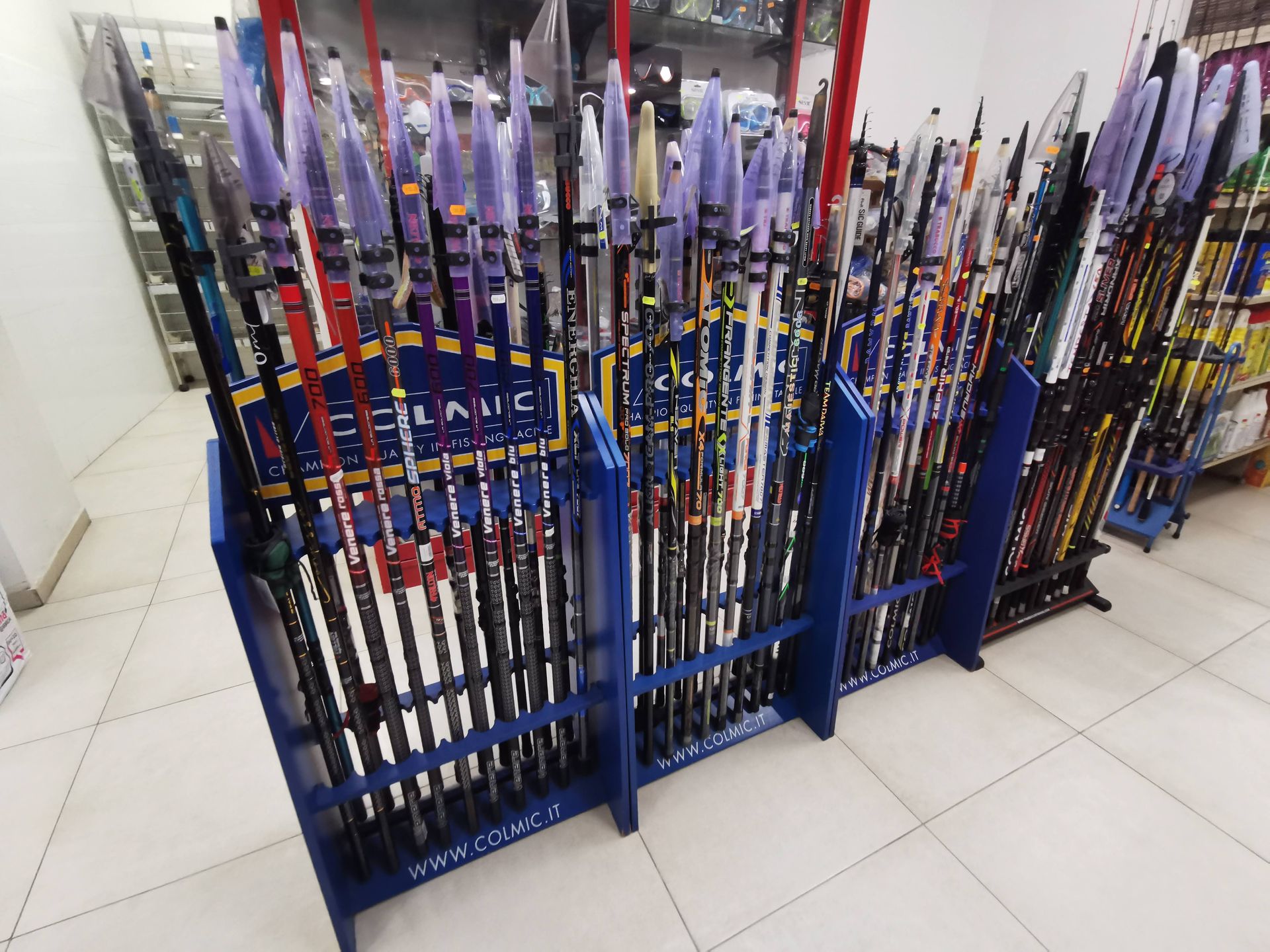 Un espositore da negozio presenta diverse canne da pesca, caratterizzate da punte protettive viola e telai in metallo blu.