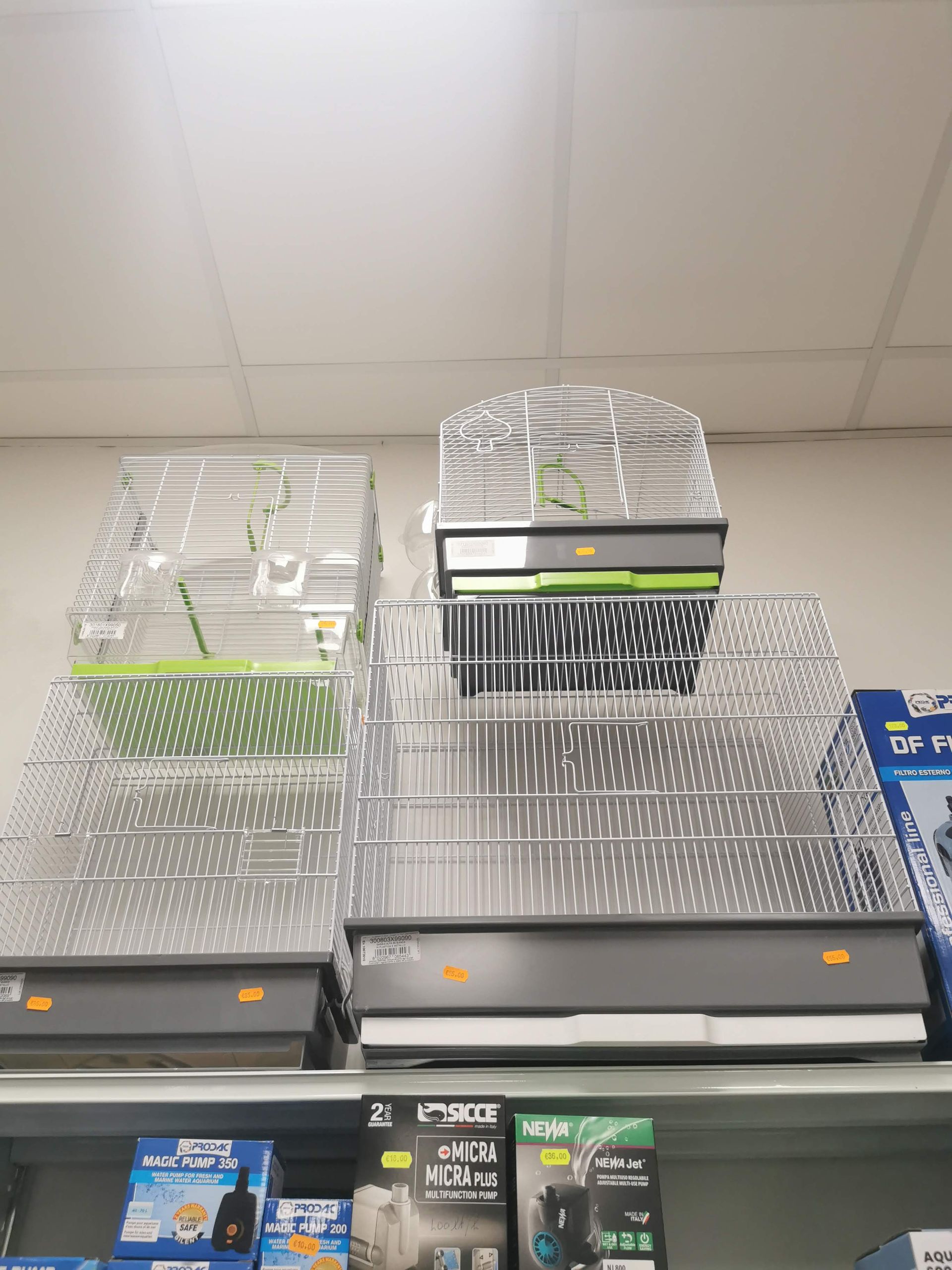 In un negozio, su uno scaffale sono esposte diverse gabbie per uccelli con basi grigie e verdi.