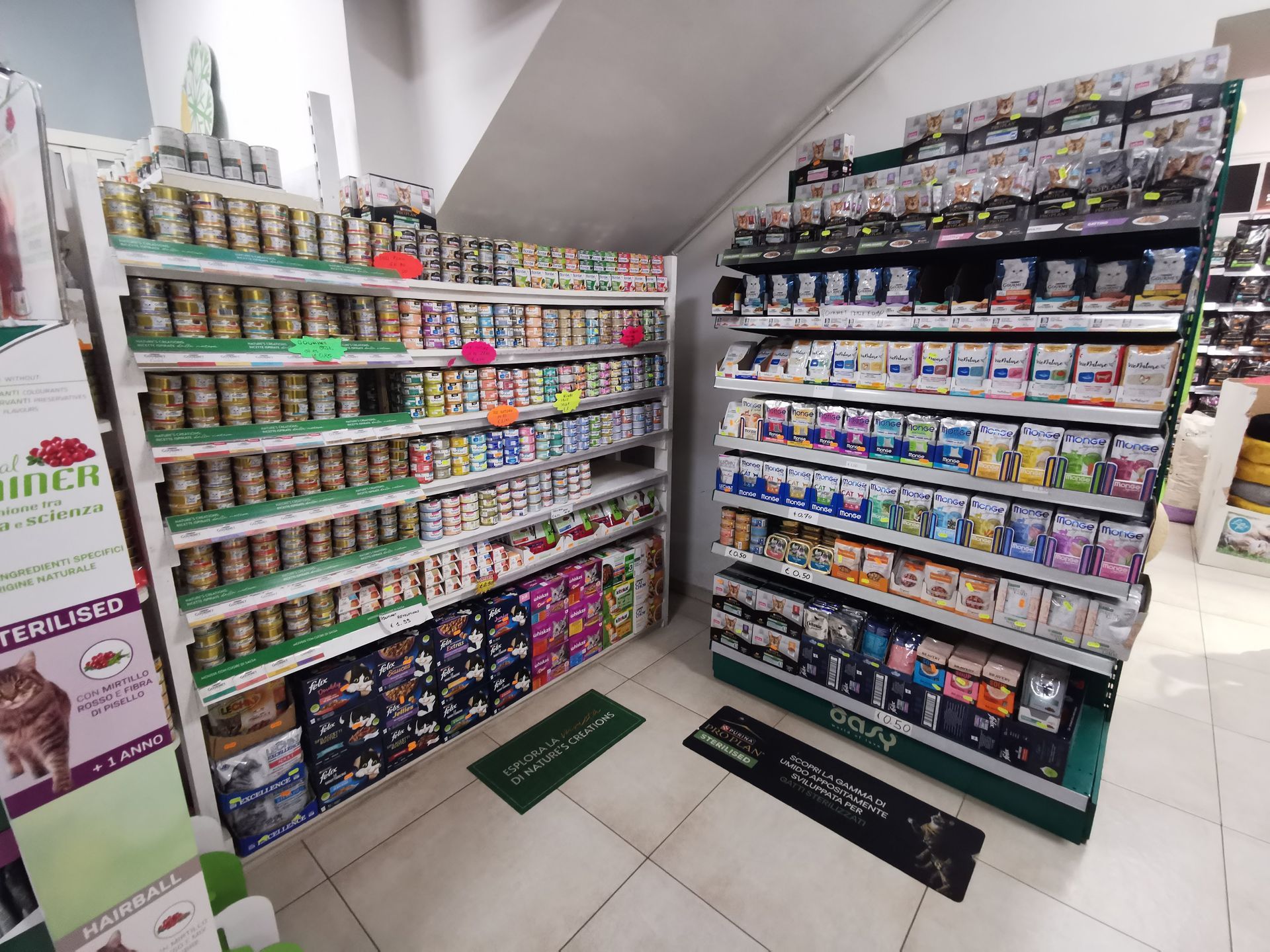 Gli scaffali di un negozio sono pieni di lattine di cibo per animali di vario tipo e piccole confezioni di accessori per animali, con due tappetini sul pavimento piastrellato.