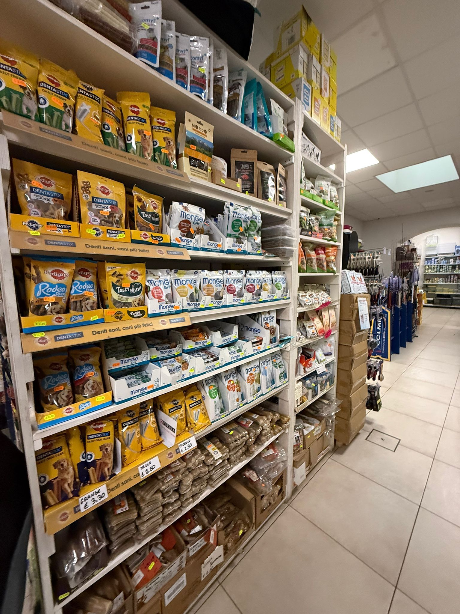 Scaffalature alte e luminose in un negozio di articoli per animali, piene di sacchi di cibo e snack di vario genere.