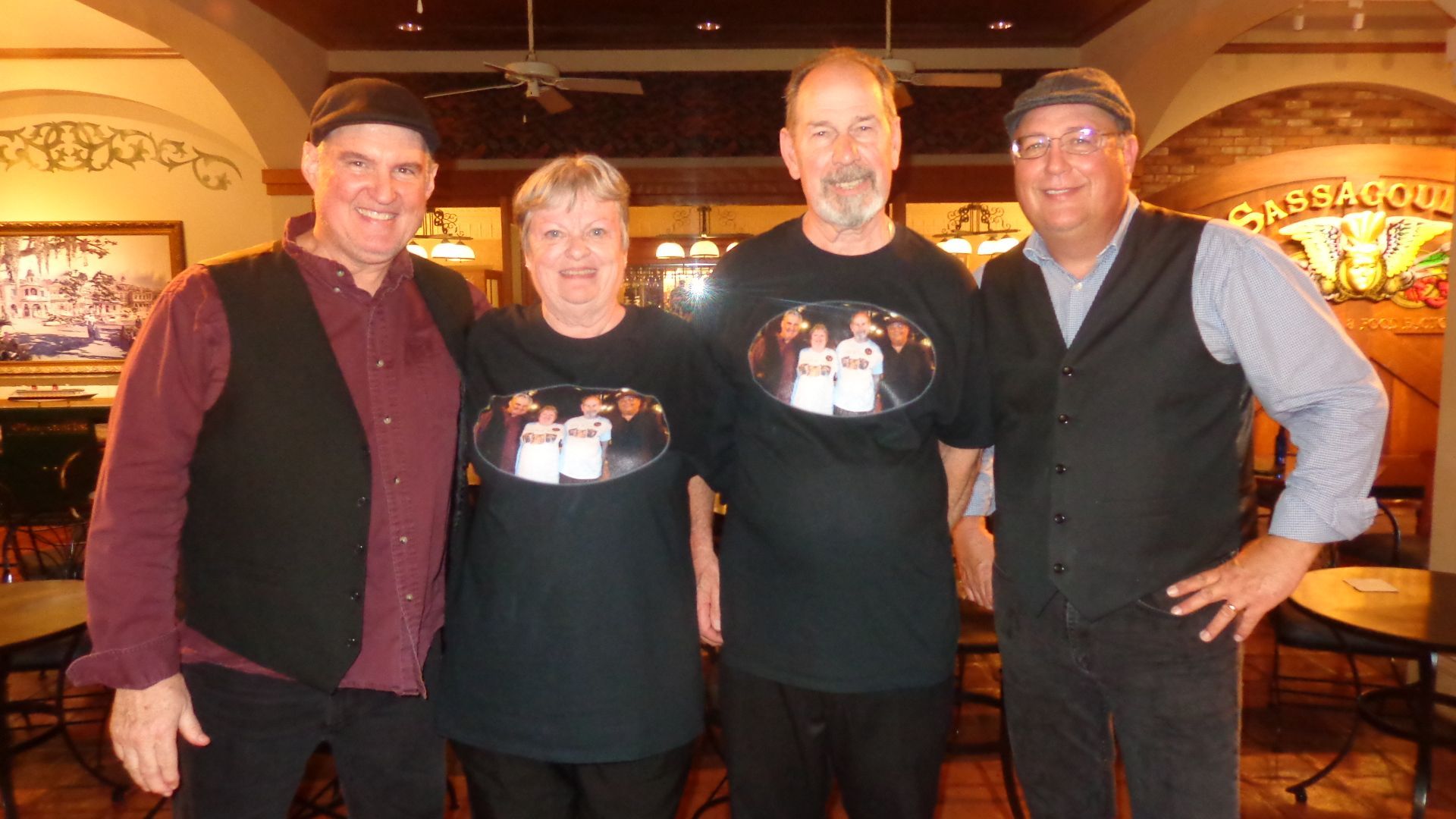 Four people in a restaurant, two wearing matching shirts with a photo. Two men wear vests. All smiling.