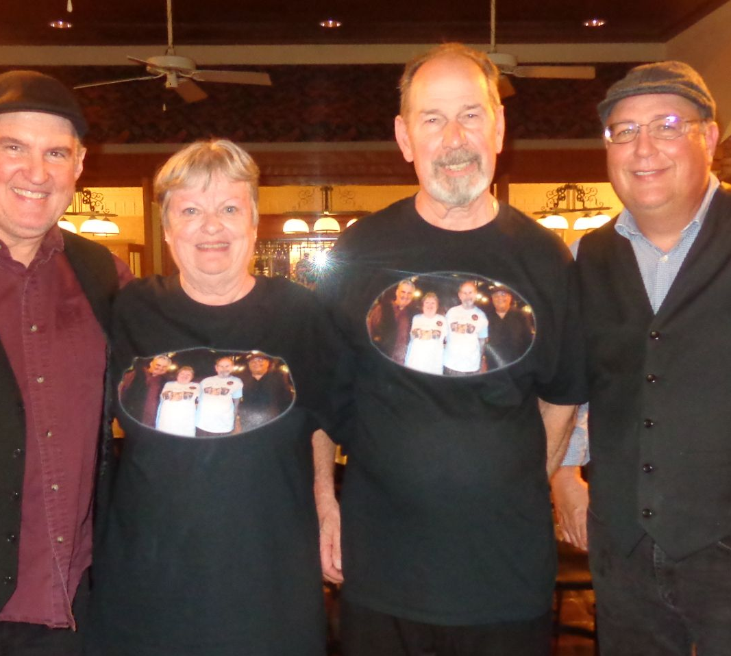 Four people, two wearing t-shirts with an image, pose indoors. Warm lighting. Smiling.