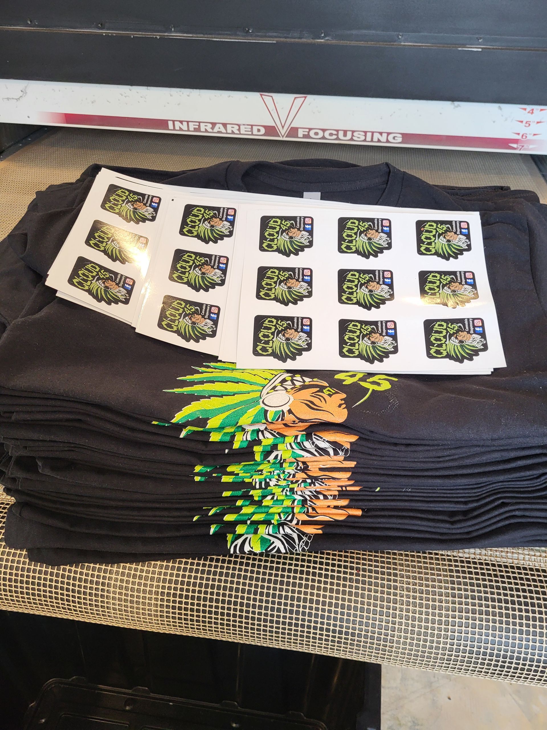 Stacks of gray t-shirts with a printed design, with sticker sheets on top, on a patterned surface.