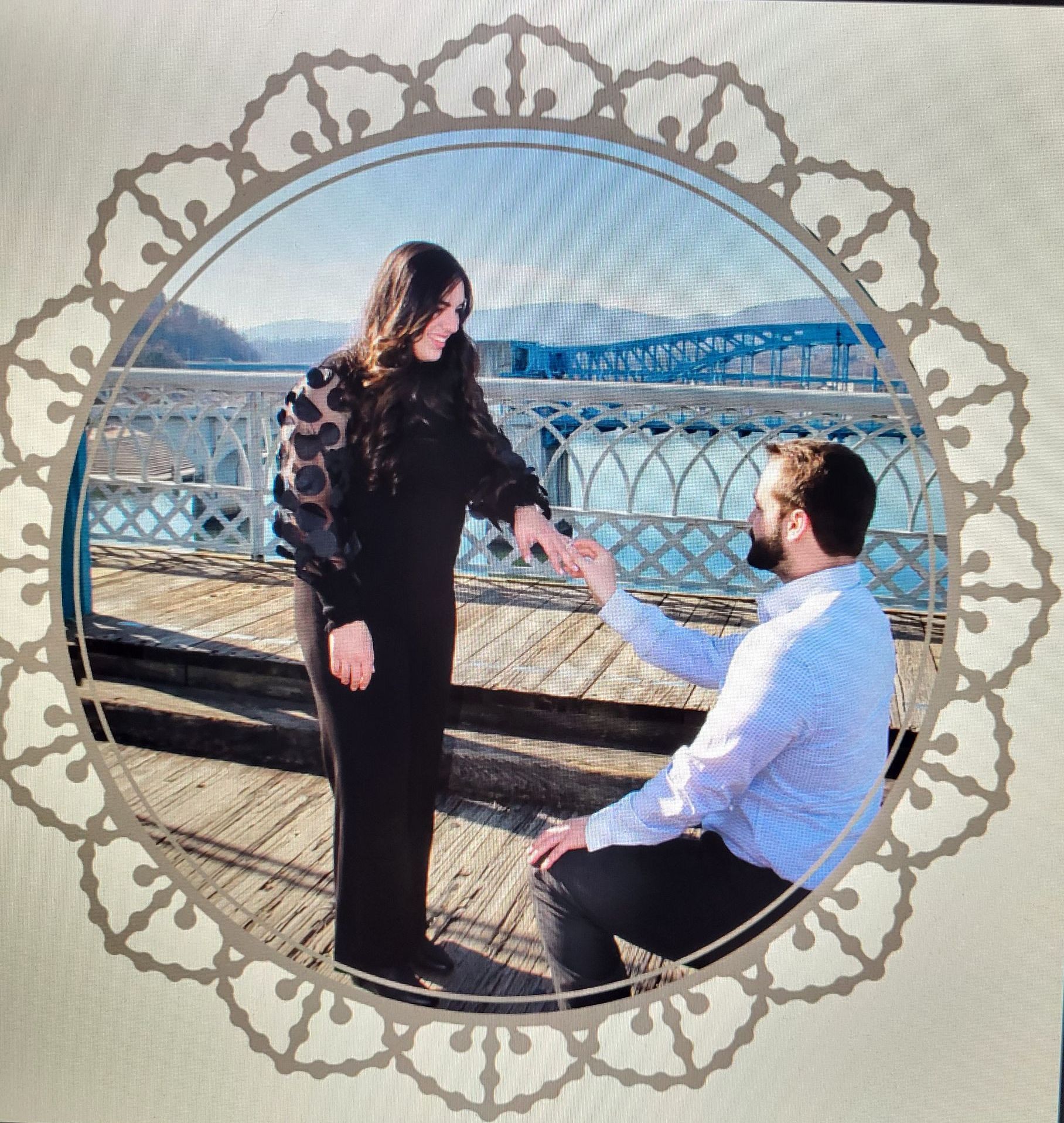 Man proposing to woman on a pier; mountain and bridge background.