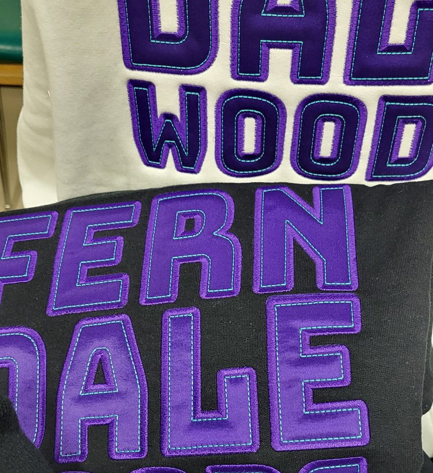 Close-up of two sweatshirts. One is white, the other black. Both have purple letters with blue stitching.