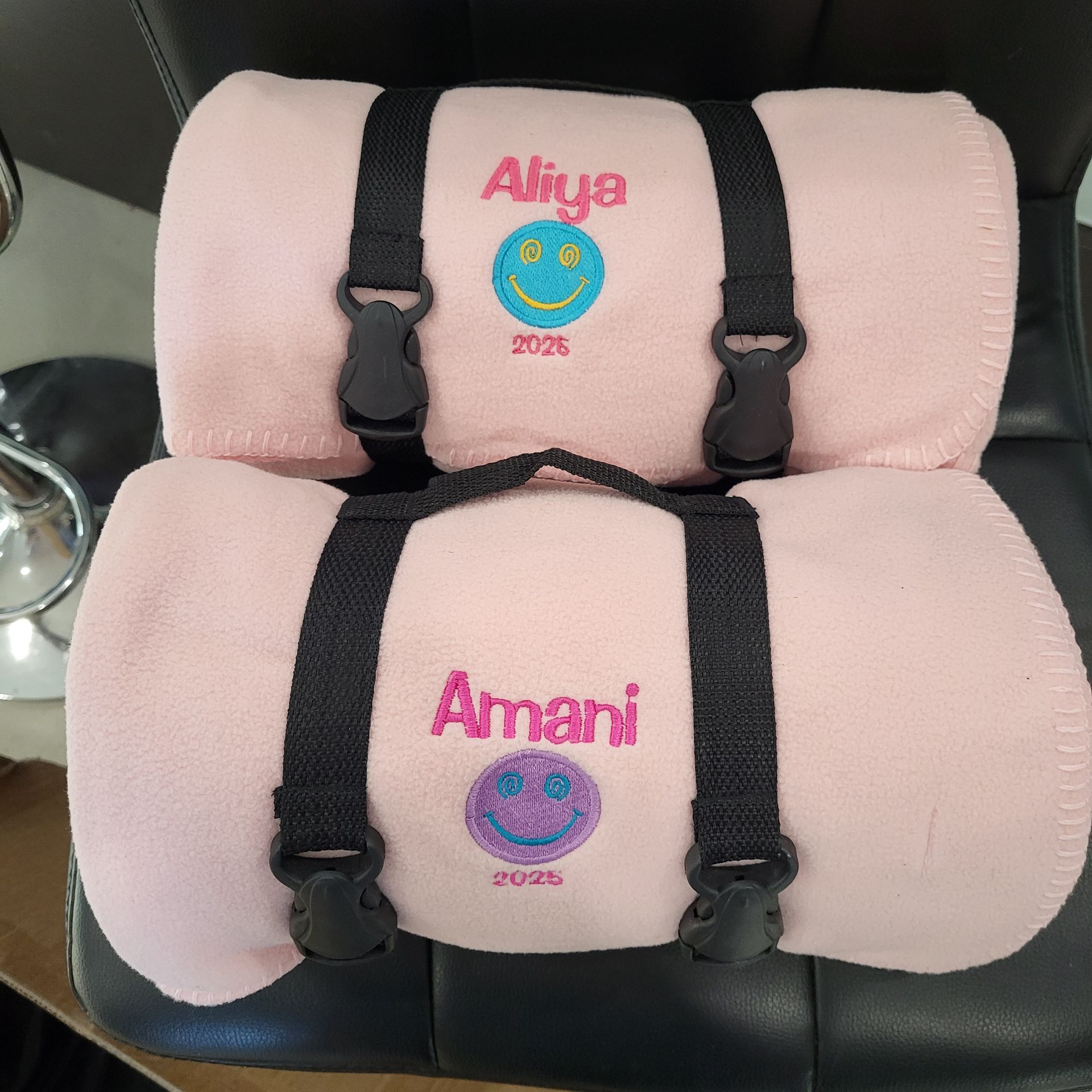 Two rolled pink blankets with black straps, each embroidered with a name, smiley face, and year.