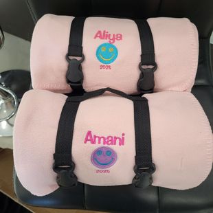 Two pink, rolled blankets with black straps and embroidered names, smiley faces, and years (2024 and 2025).